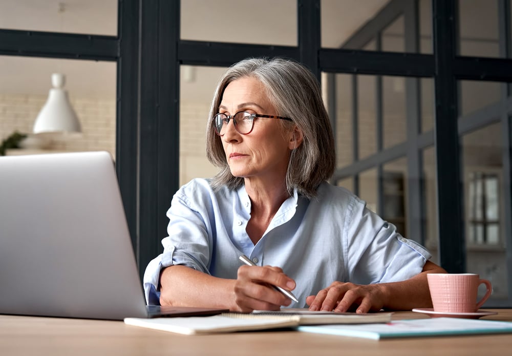 Woman reviewing W-8 BEN forms and taking notes while referencing records on her laptop. 
