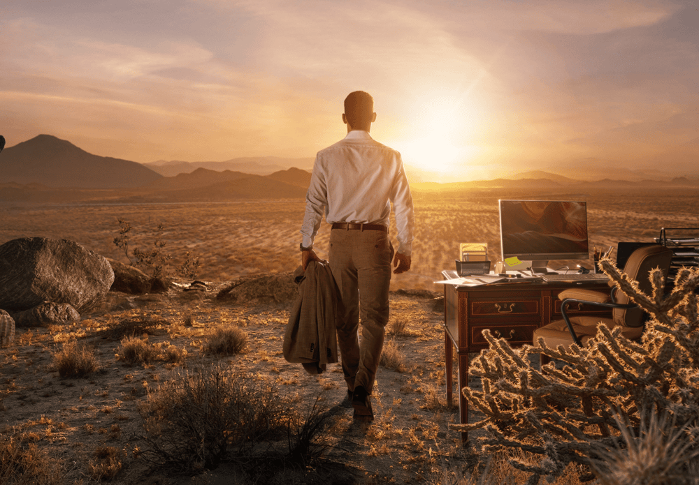 Man standing in a desert landscape walking toward a remote outdoor workspace with a desk, computer, and office chair at sunrise.