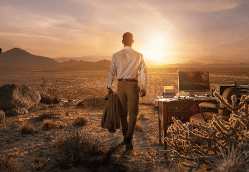 Man standing in a desert landscape walking toward a remote outdoor workspace with a desk, computer, and office chair at sunrise.