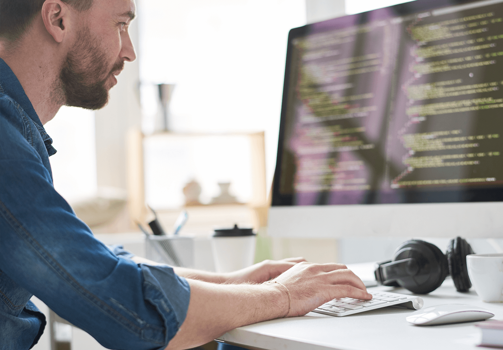A person coding on a computer with headphones on the desk