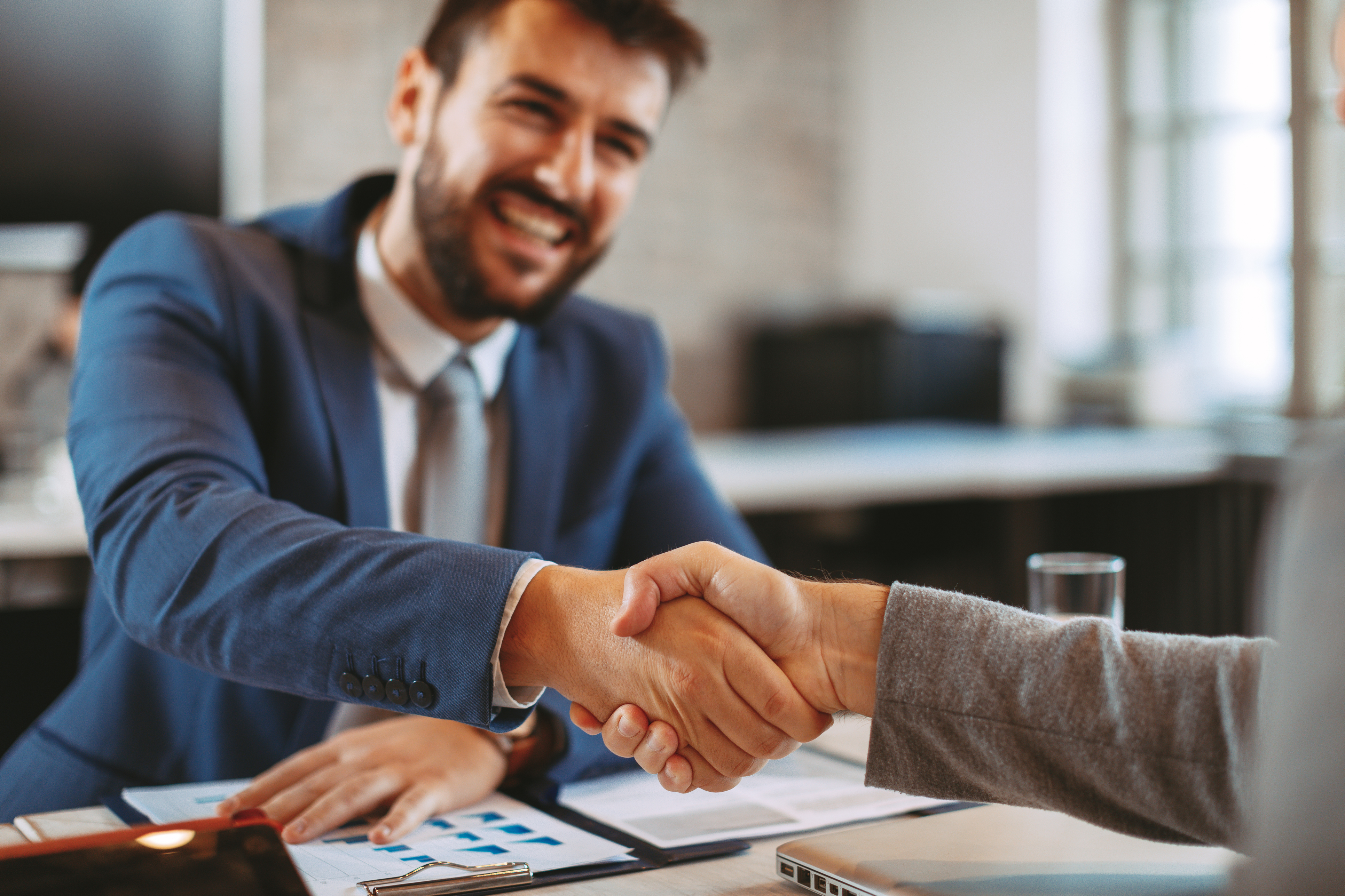 A close-up of a smiling man in a suit shaking hands with a colleague over a desk with business documents in a bright office.